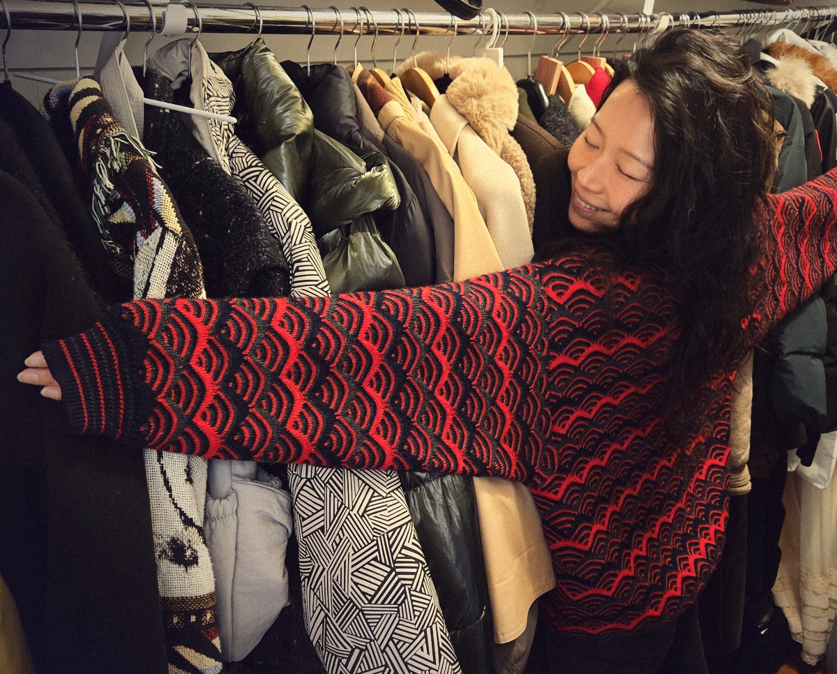 Christina hugging a rack of her coats, arms spread wide, in a red and navy wave-patterned sweater